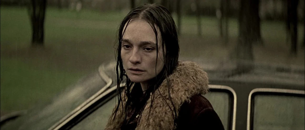 Dark Water (2005) movie still: medium — A woman stands in the rain in a wooded park area behind a vehicle. She wears a coat with a faux-fur collar…