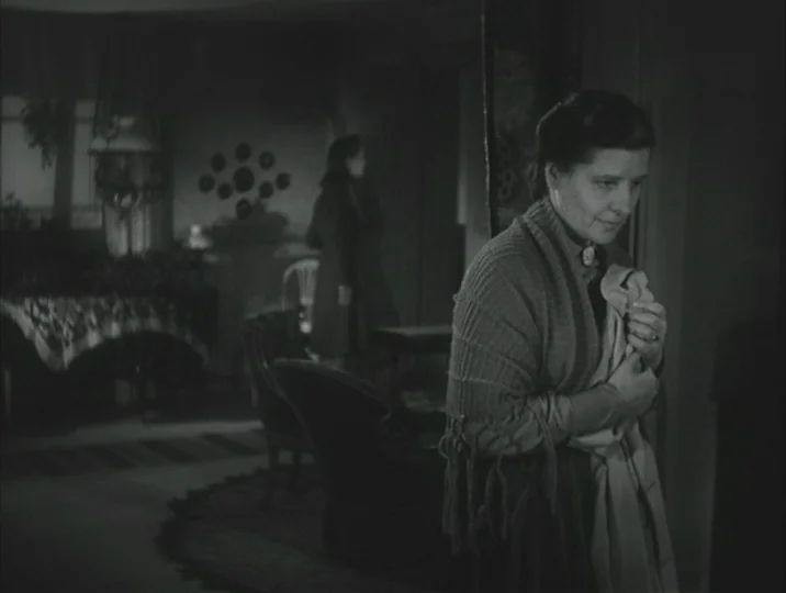 Crisis (1946) movie still: medium — A dimly lit room with an arrangement of period furniture and a decorative wall clock. A woman stands in the…