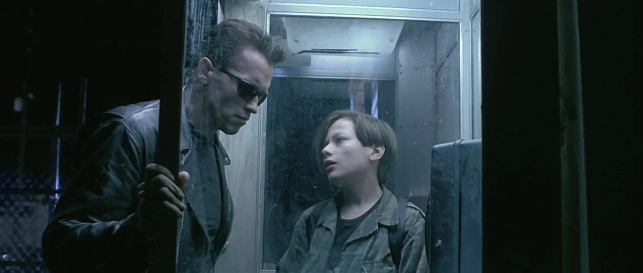 Terminator 2: Judgement Day (1991) movie still: medium — A man and a teenager stand inside a dimly lit outdoor glass booth at night. The man wearing…