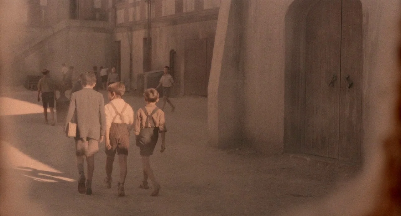 The Devils Backbone (2001) movie still: wide — A dusty school courtyard with light stone walls and large arched wooden doors. Three young boys walk…