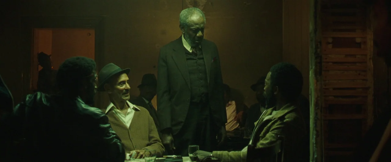 Mangrove (2020) movie still: medium — A dimly lit room with peeling walls and wooden trim houses several seated men. An elderly man in a suit stands…