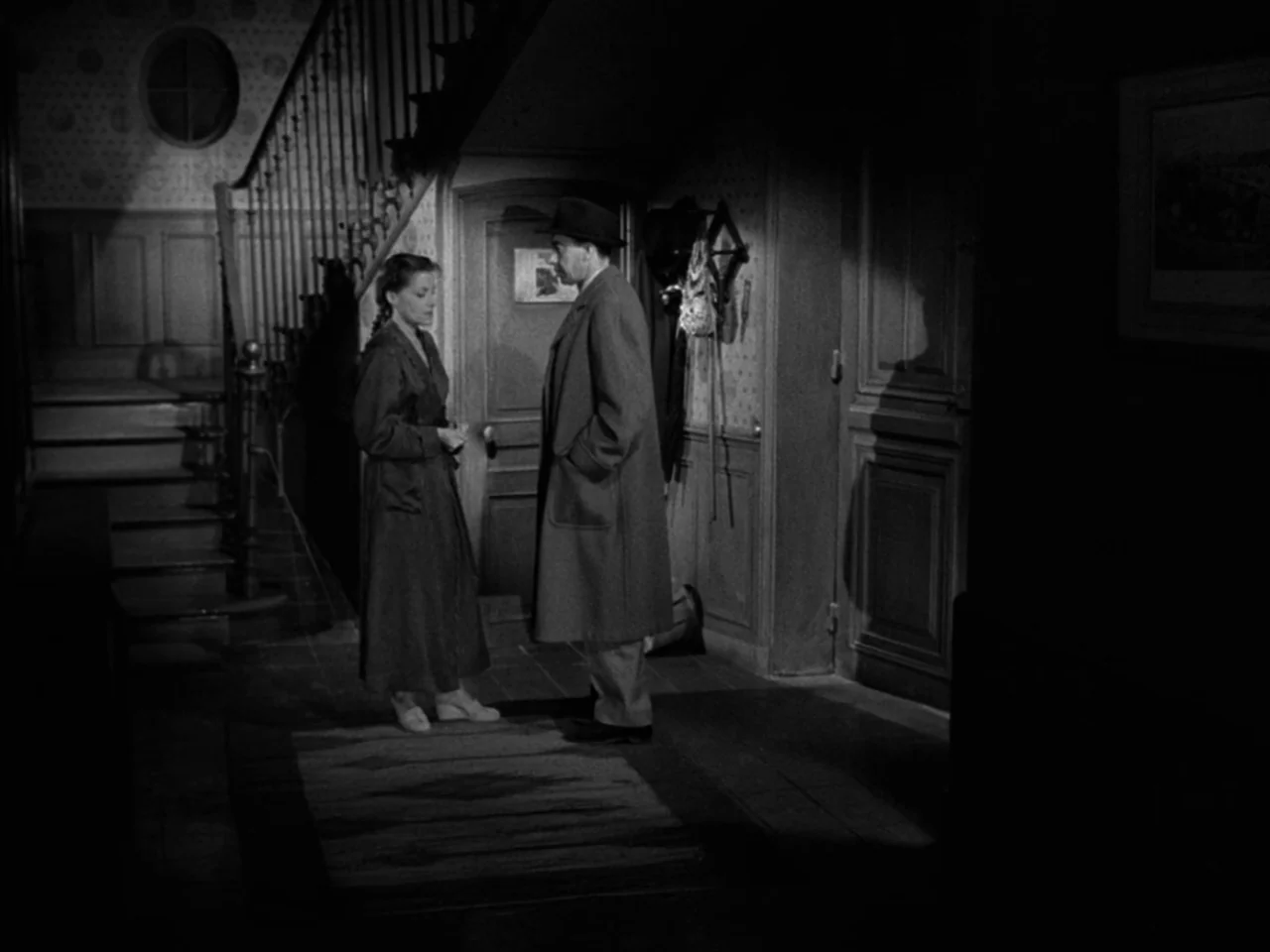 Diabolique (1955) movie still: fullbody — A dark foyer with a wooden staircase and floral patterned wallpaper. A man and a woman stand on a rug in…