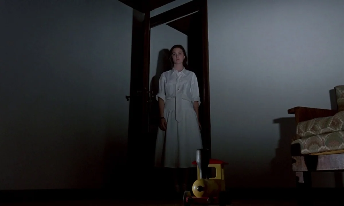 Phenomena (1985) movie still: fullbody — A dimly lit interior with stark white walls and a dark open doorway. A young woman in a white outfit stands…