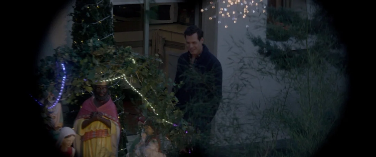 Elle (2016) movie still: medium — A man stands outdoors near a small nativity scene illuminated by decorative lights. The scene is viewed through a…