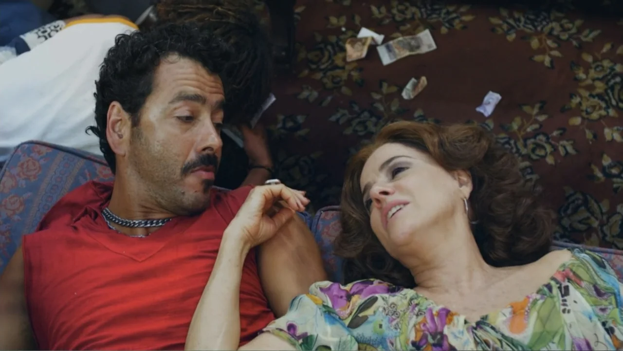 For Sale or Rent (2013) movie still: medium — A domestic living room space with a patterned rug on the floor. A man and a woman lie together on a…