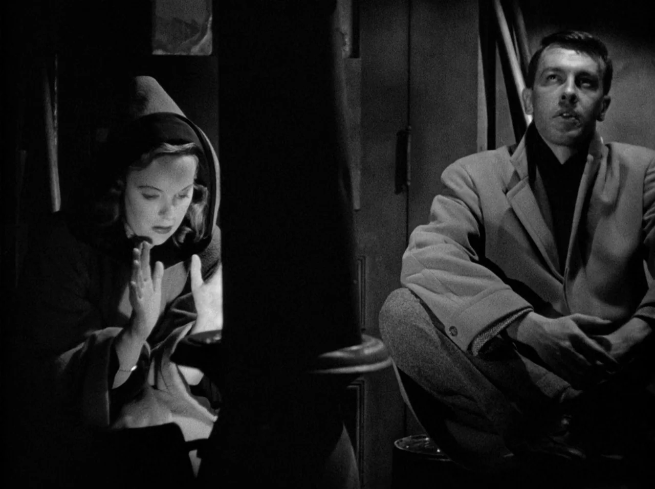 Gun Crazy (1950) movie still: medium — A dark, dimly lit room contains a central stove dividing two people. The woman warms her hands near the light…