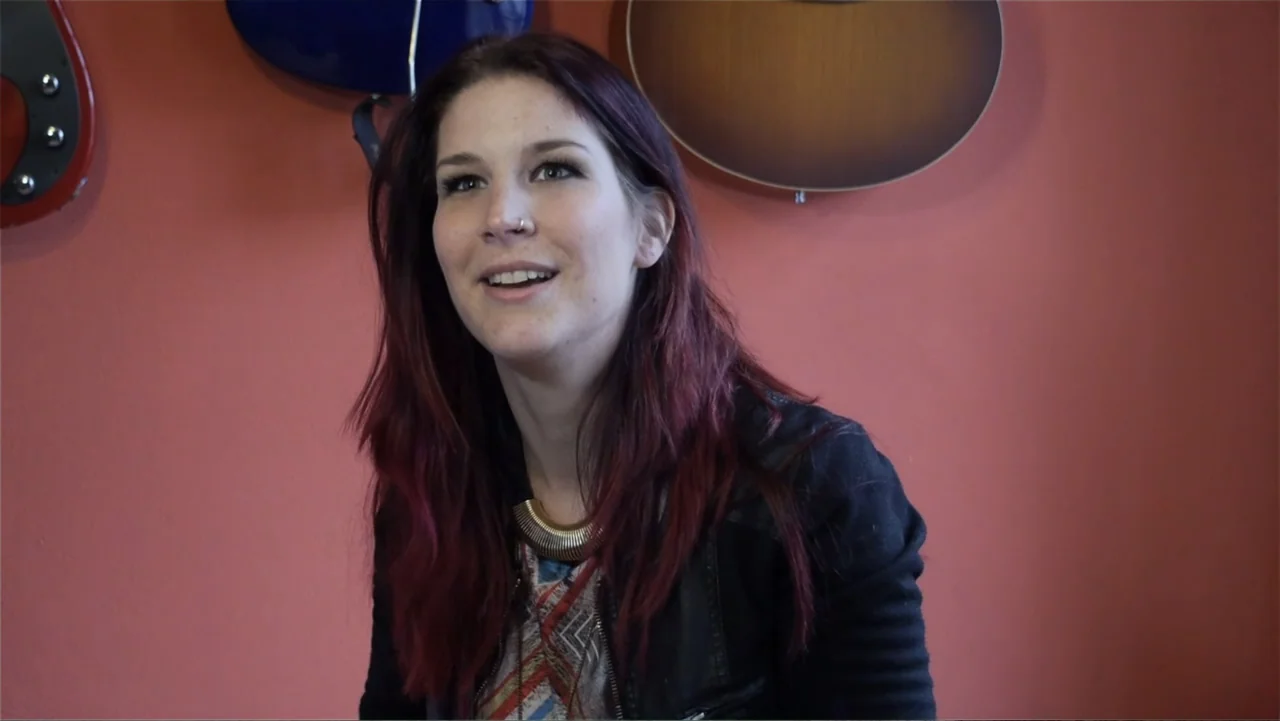 Soaring Highs and Brutal Lows: The Voices of Women in Metal (2015) movie still: medium — A room with a solid muted orange wall and guitars mounted…
