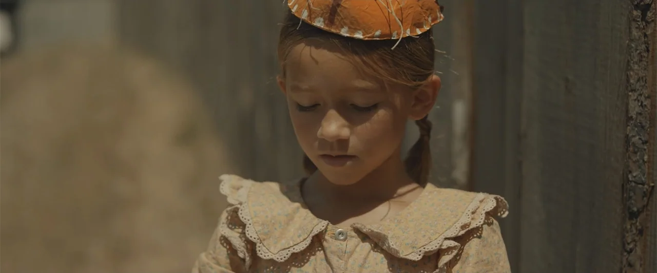 Ghosts of the Ozarks (2022) movie still: closeup — A young girl stands in front of a rustic wooden fence on a bright day. She wears a floral-print…