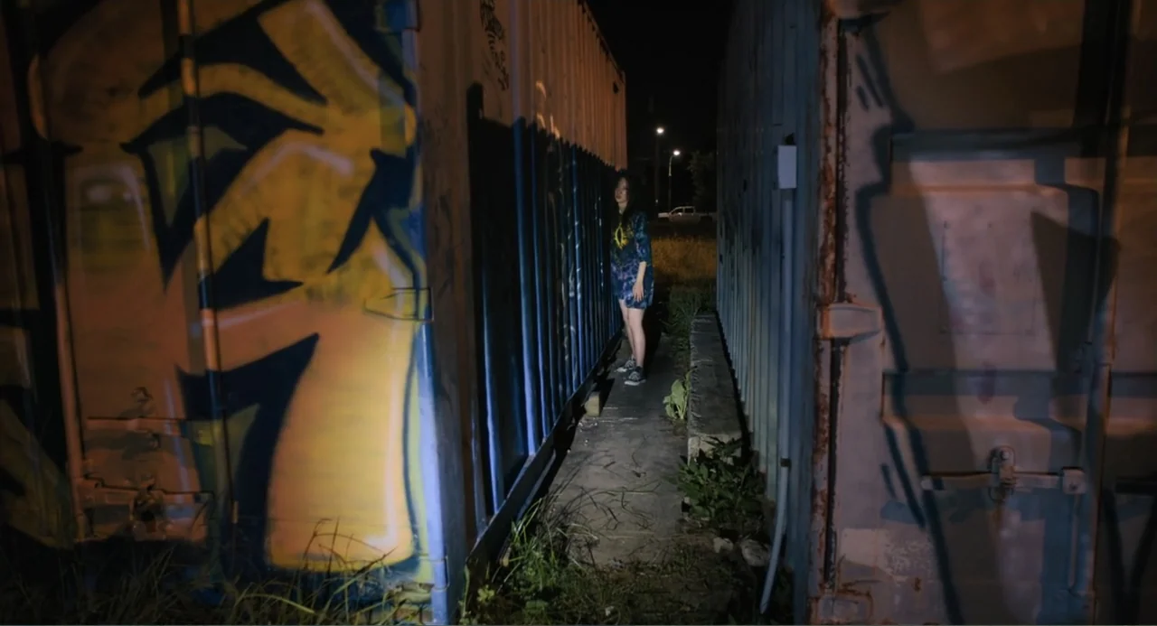 Mona Lisa and the Blood Moon (2022) movie still: fullbody — A dark, narrow outdoor corridor is flanked by two large, industrial shipping containers.…