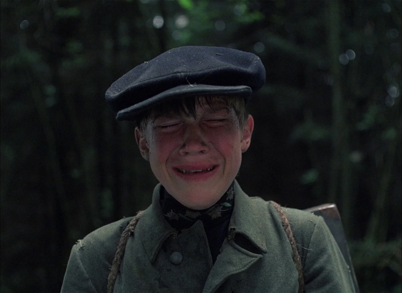 Come and See (1985) movie still: closeup — A dense, dark forest filled with blurred trunks and green foliage surrounds the boy. A young male in a…