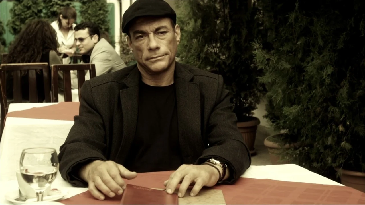 Assassination Games (2011) movie still: medium — An outdoor restaurant terrace features a table with a white cloth and a red runner. A man in a dark…