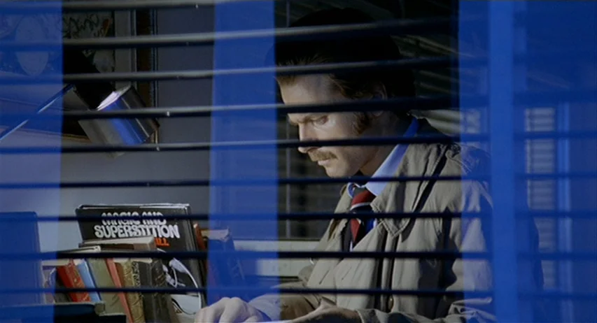 The Fifth Cord (1971) movie still: medium — An office interior is obscured by horizontal blue blinds in the foreground. A man in a trench coat…