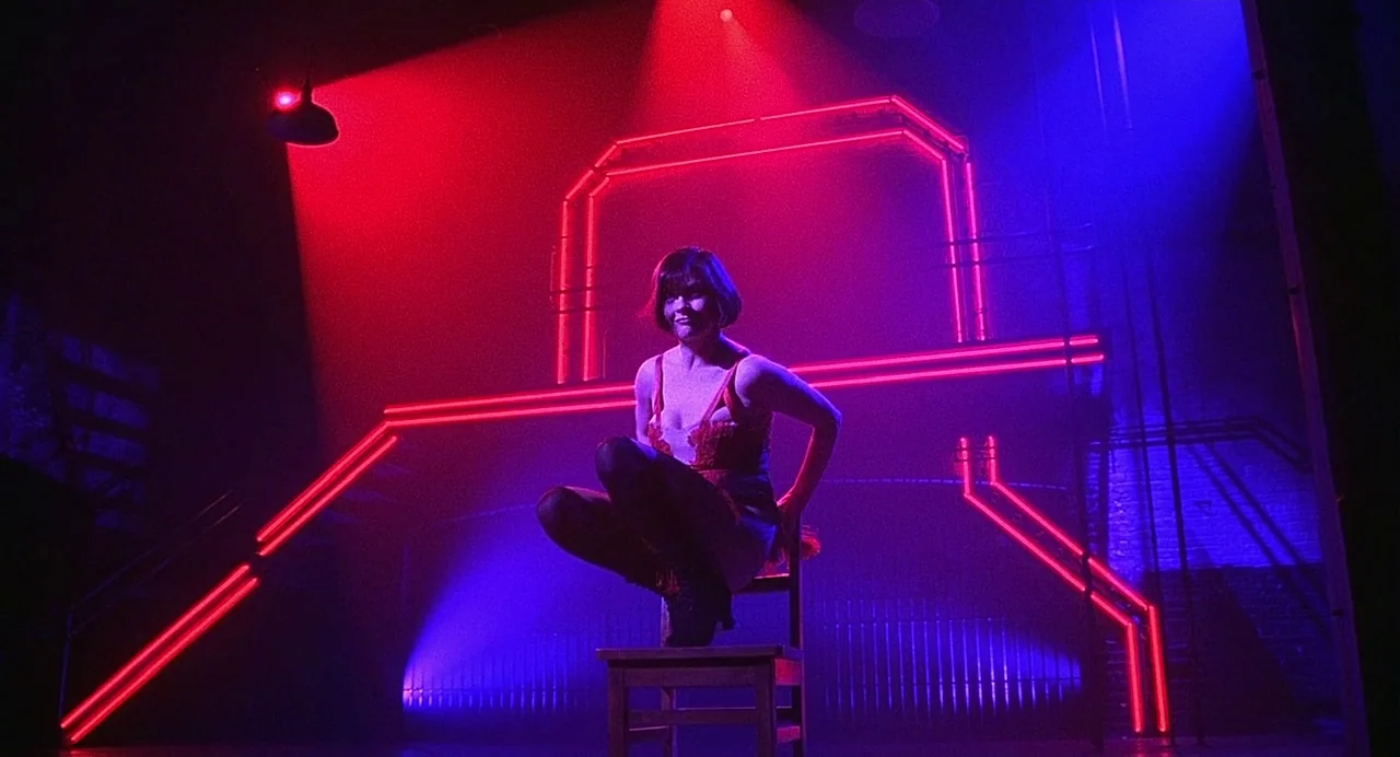 Chicago (2002) movie still: fullbody — A theatrical stage environment features a dark brick wall illuminated by geometric neon tubing. A woman sits…