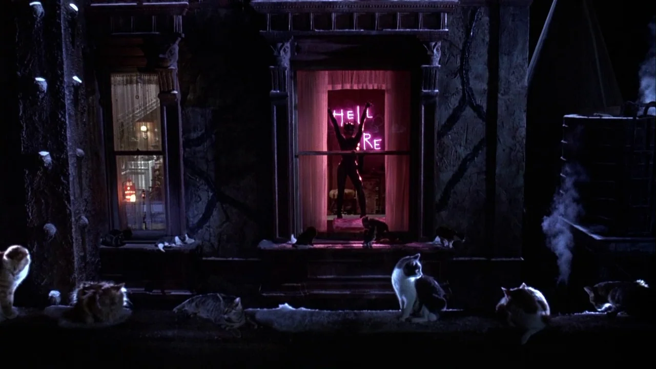 Batman Returns (1992) movie still: wide — A dark urban building exterior features a central glowing pink neon sign. A female figure in a tight black…