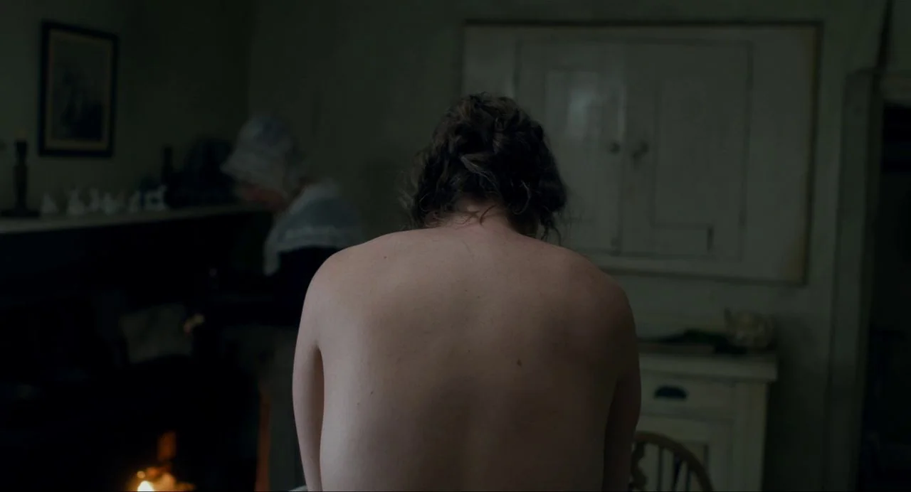 Ammonite (2020) movie still: medium — A dimly lit 19th-century cottage interior features a bare-backed woman seated in the foreground. Another woman…