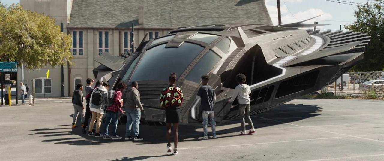 Black Panther (2018) movie still: wide — A group of teenagers stands in an asphalt parking lot surrounding a landed spaceship. The metallic craft is…