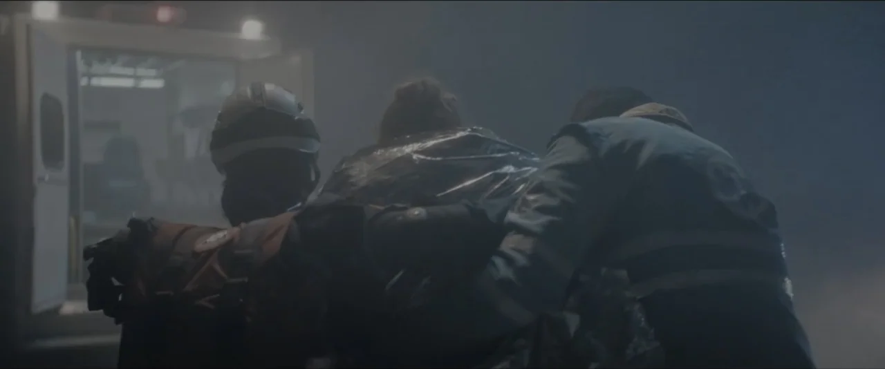 Quicksand (2023) movie still: medium — A dark, foggy outdoor space features a parked ambulance with its rear doors standing open. Two paramedics…