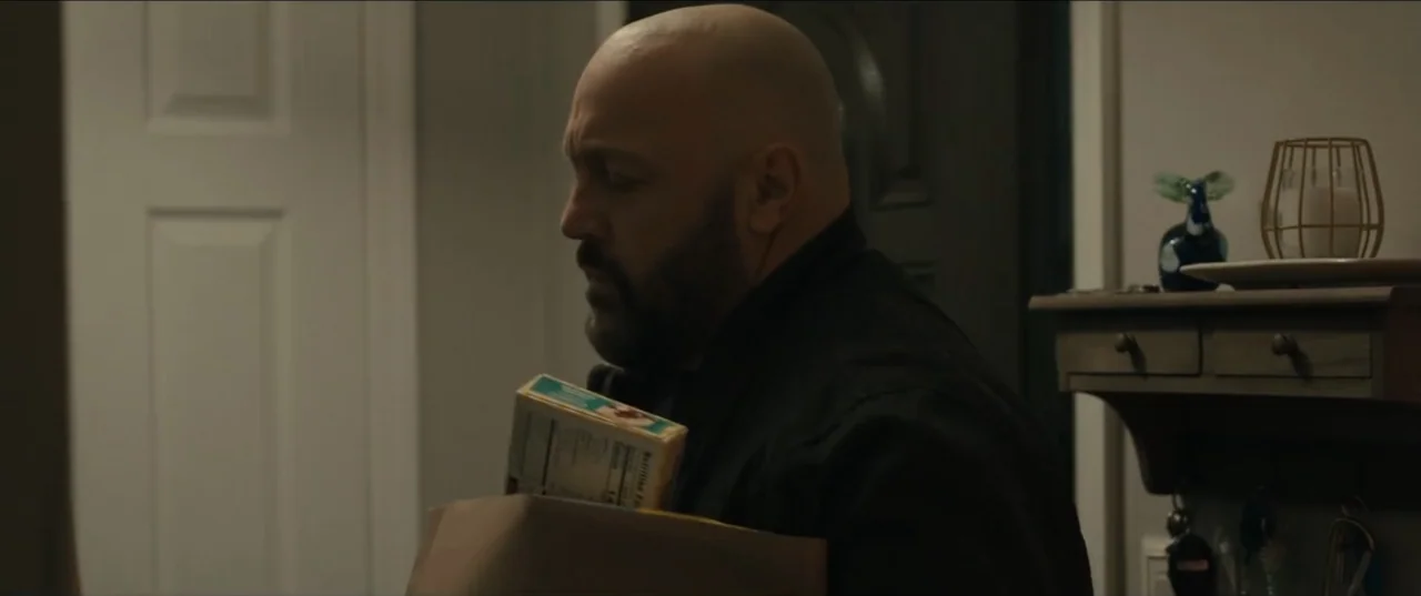 Guns Up (2025) movie still: medium — A dimly lit interior hallway features a closed door and a small wall shelf. A bald man with a dark beard holds…