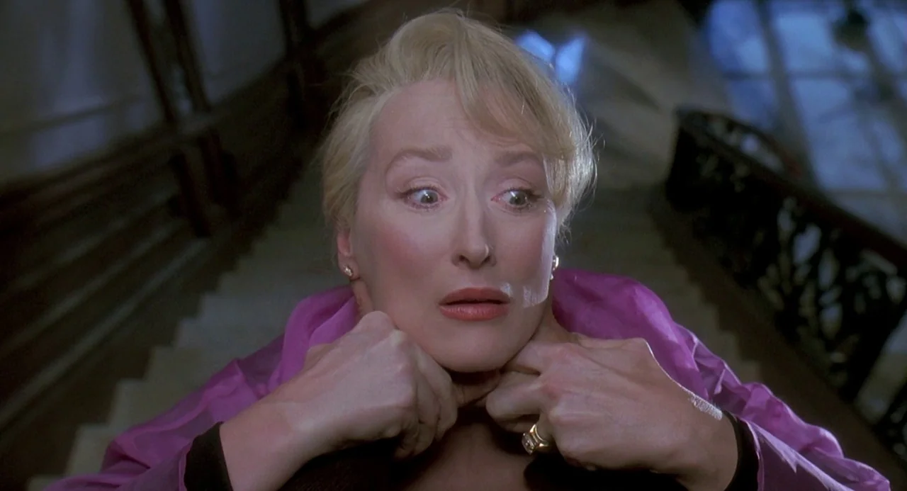Death Becomes Her (1992) movie still: closeup — A woman stands at the top of a dimly lit, shadowed residential staircase at night. She clutches her…
