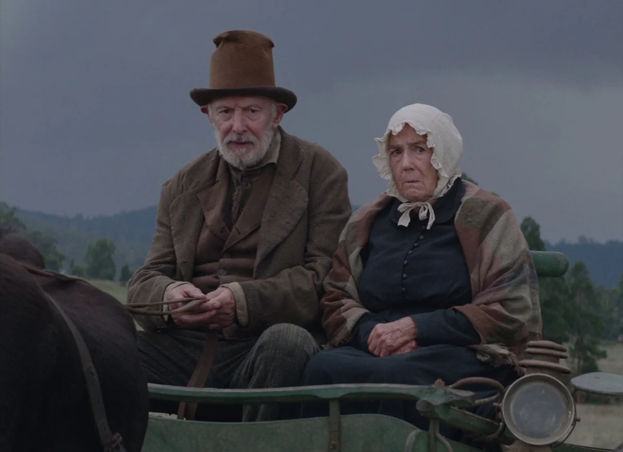 The Nightingale (2018) movie still: medium — An elderly couple sits side-by-side on a wooden cart against a dark, stormy sky. The man holds leather…