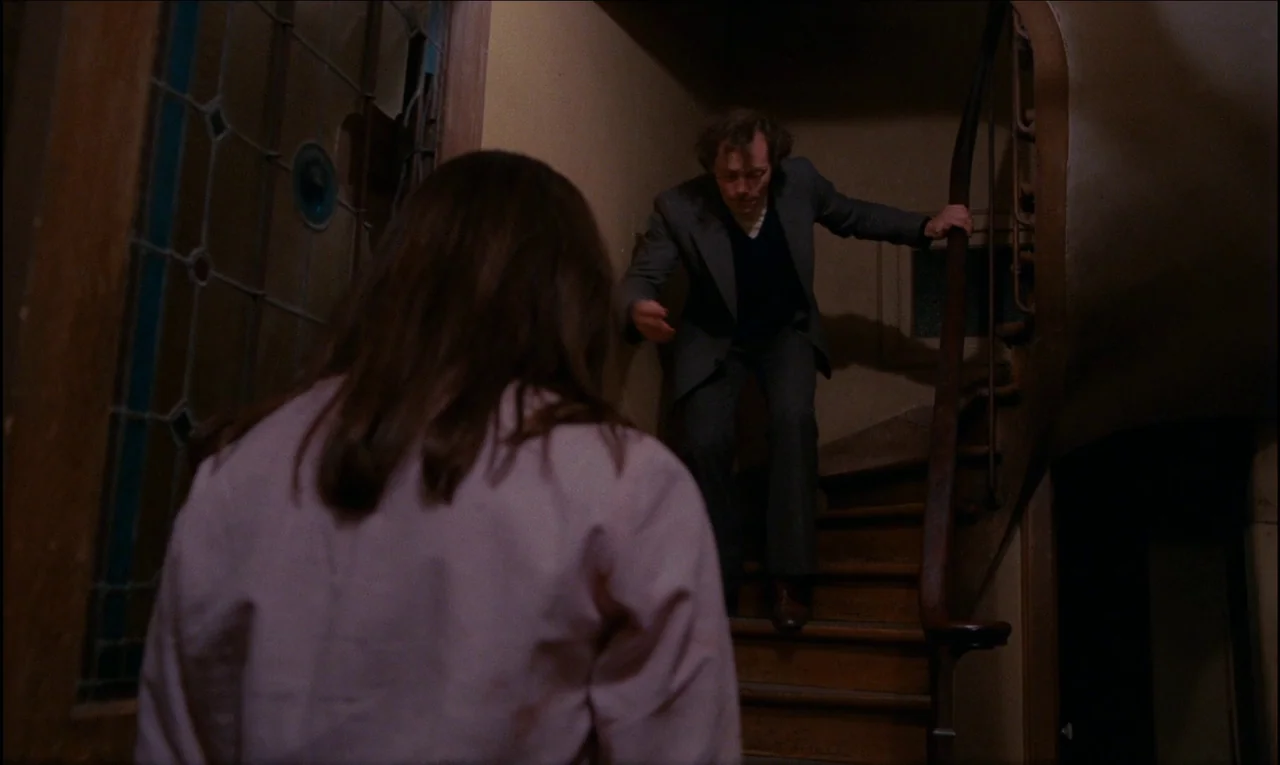 Serie Noire (1979) movie still: medium — An aged interior staircase with a stained-glass window and wooden banister is dimly lit. A man in a suit…