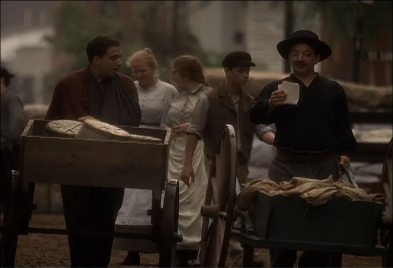 Vendetta (2022) movie still: medium — A group of people dressed in early twentieth-century clothing walks past several wooden pushcarts. One man…