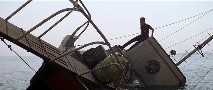 Jaws movie still: wide — A partially submerged boat tilts heavily into the water under an overcast sky. A man sits calmly on the exposed side of the…