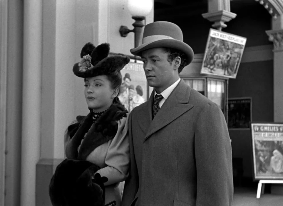 The Magnificent Ambersons (1942) movie still: medium — A man and a woman in 1900s formal attire stand near a building entrance. The couple walks…