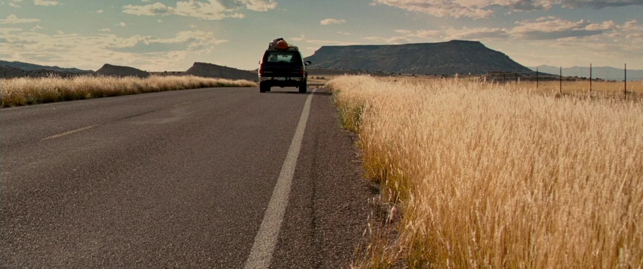Carriers (2009) movie still: wide — A rural highway stretches through a dry grassland valley under a partially clouded sky. A small black car with…