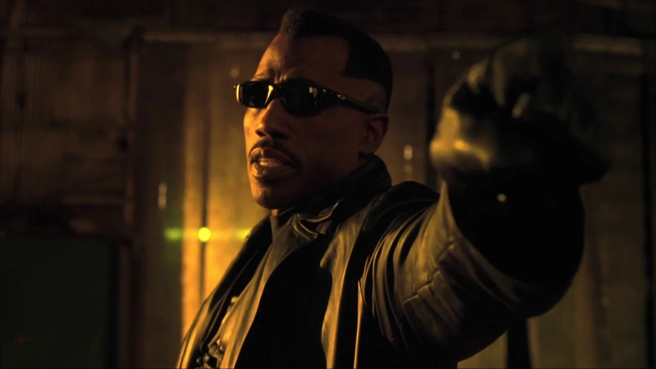Blade II (2002) movie still: closeup — An dimly lit interior with dark textured walls illuminated by a warm glow. A man in a leather coat holds his…