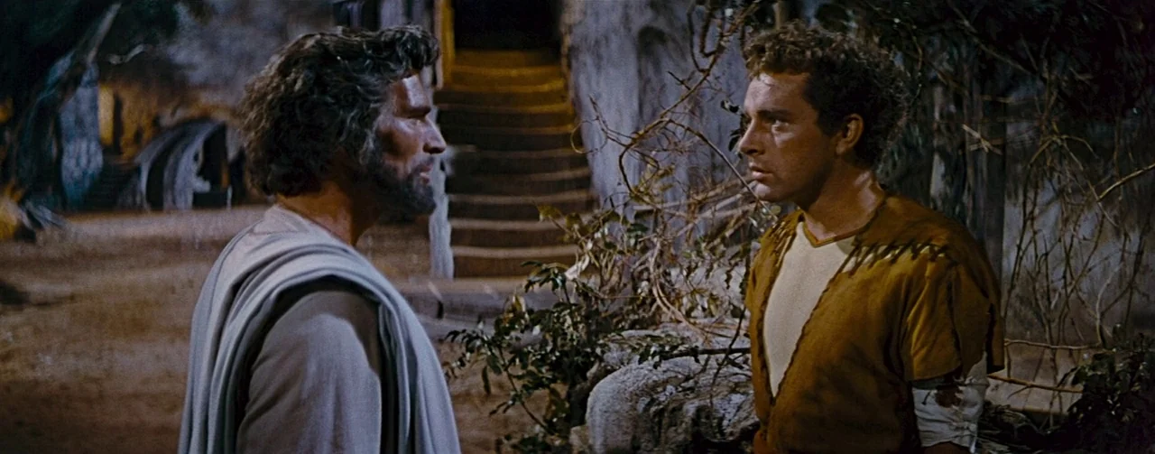 The Robe (1953) movie still: medium — A dark outdoor space at night with bare branches and earthen textures. Two men in period attire stand facing…