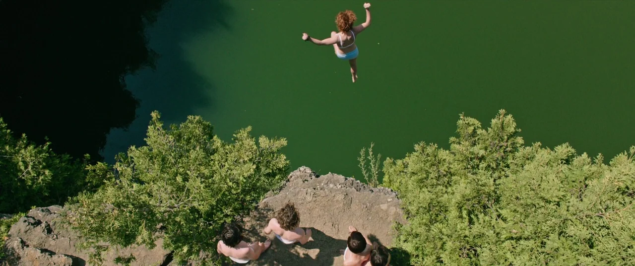 It (2017) movie still: wide — A group of people sits on a limestone ledge overlooking a murky green body of water. A girl mid-air jumps from the…