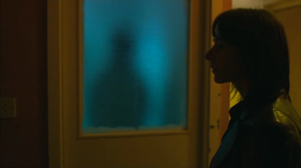 Red Road (2006) movie still: medium — A dim indoor space features a wall with a prominent frosted glass pane door. A woman stands in profile looking…