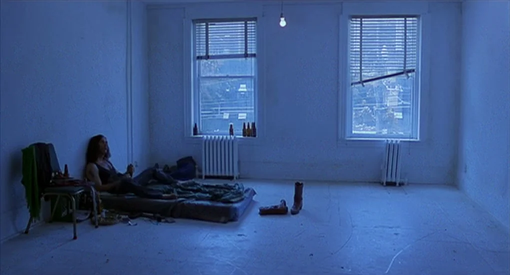 Exotica (1994) movie still: wide — A sparsely furnished room features two windows and a bare lightbulb hanging from above. A man sits alone on a low…