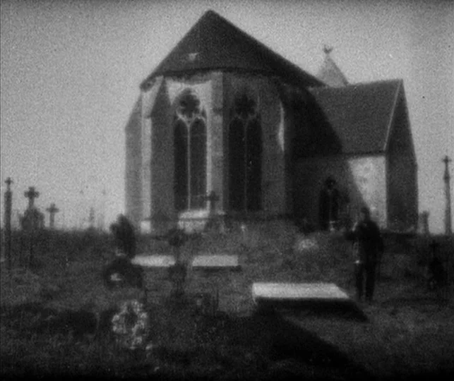 Vampyr (1932) movie still: wide — An old stone church stands in a graveyard filled with iron grave crosses. Overcast day light illuminates the grass…