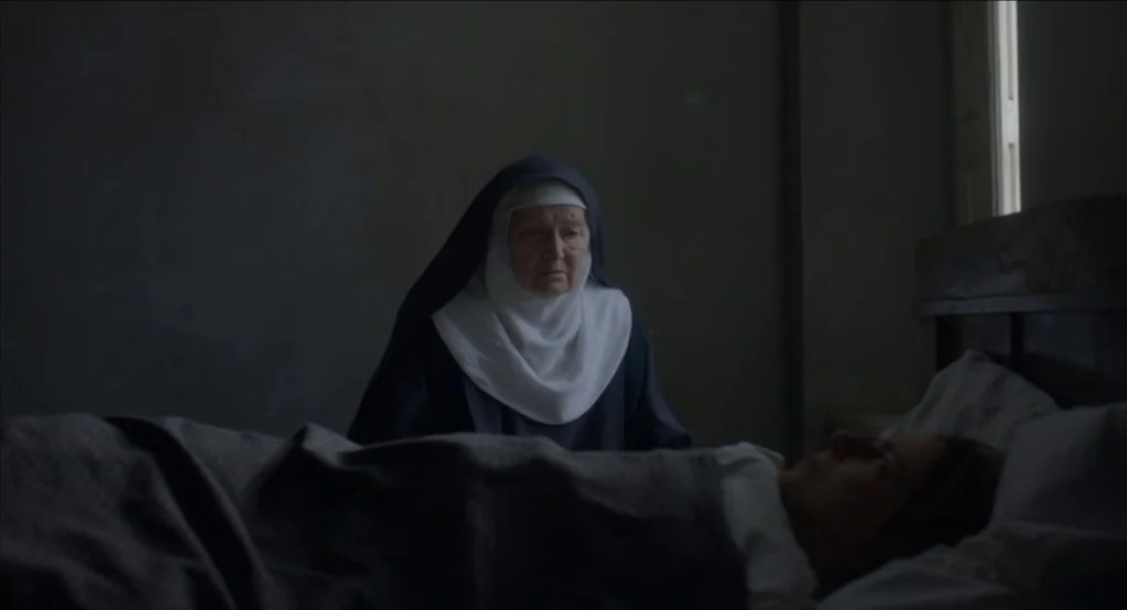 The Innocents (2016) movie still: medium — A dimly lit room with a person lying in a bed. An elderly nun wearing a habit sits watching the patient.