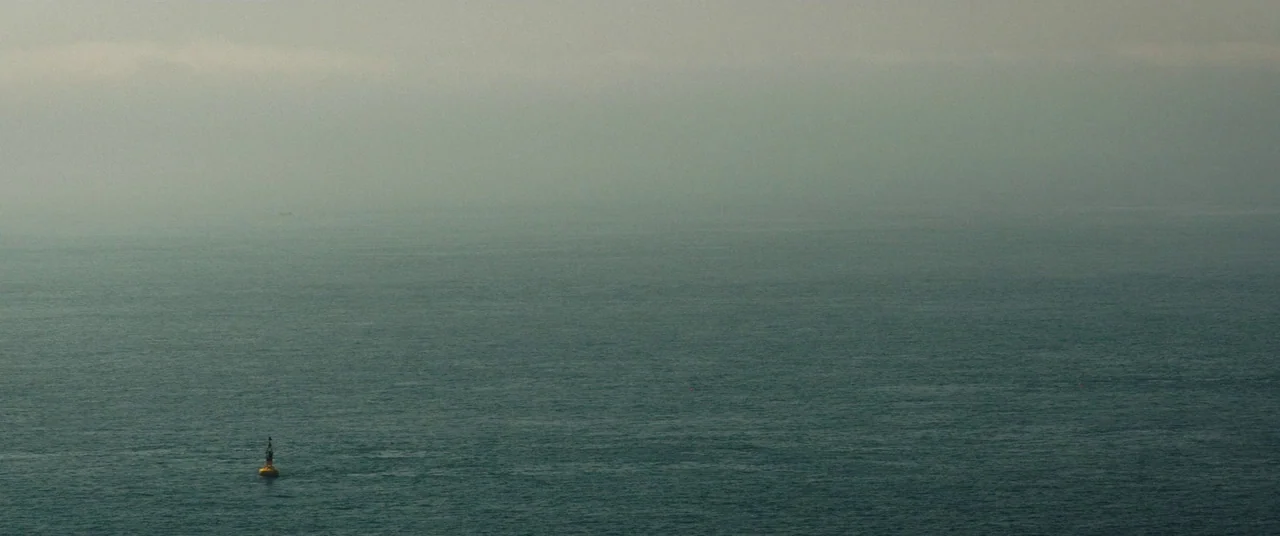Beast (2017) movie still: establishing — An expanse of greyish-blue water stretches toward a foggy, overcast horizon line. A lone yellow…