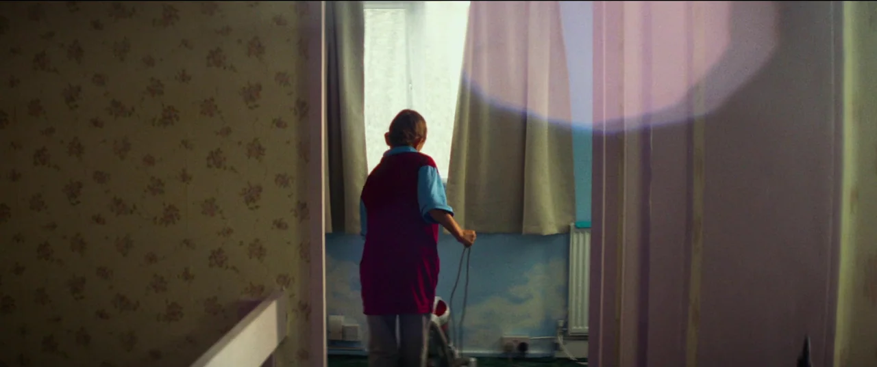 Scrapper (2023) movie still: medium — A child's room has floral wallpaper and a wall painted with a sky mural. A woman standing with her back to the…