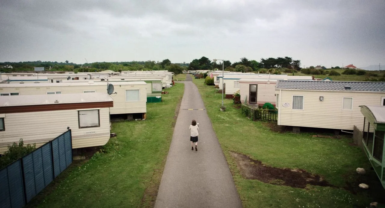 Dating Amber movie still: wide — An extensive mobile home park consists of numerous beige rectangular dwellings on grass. A lone woman walks away…