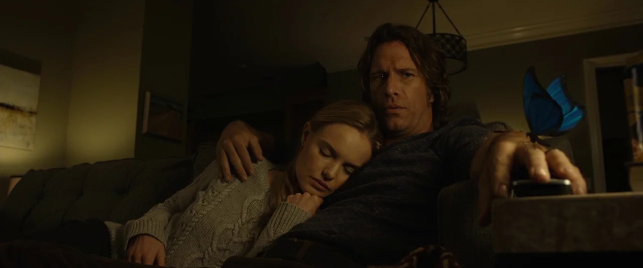 Before I Wake (2016) movie still: medium — A dark living room with a grey tufted sofa and muted wall decor. A man sits with a woman resting on his…