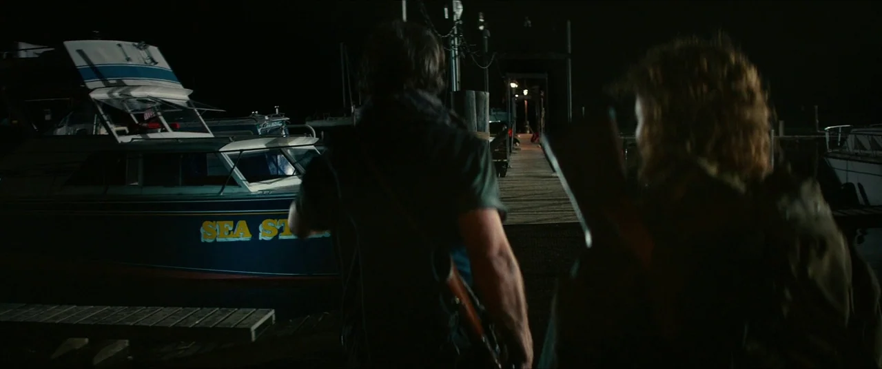 A Quiet Place Part II (2020) movie still: medium — A wooden pier at night with boats moored in the background. Two individuals walk away from the…