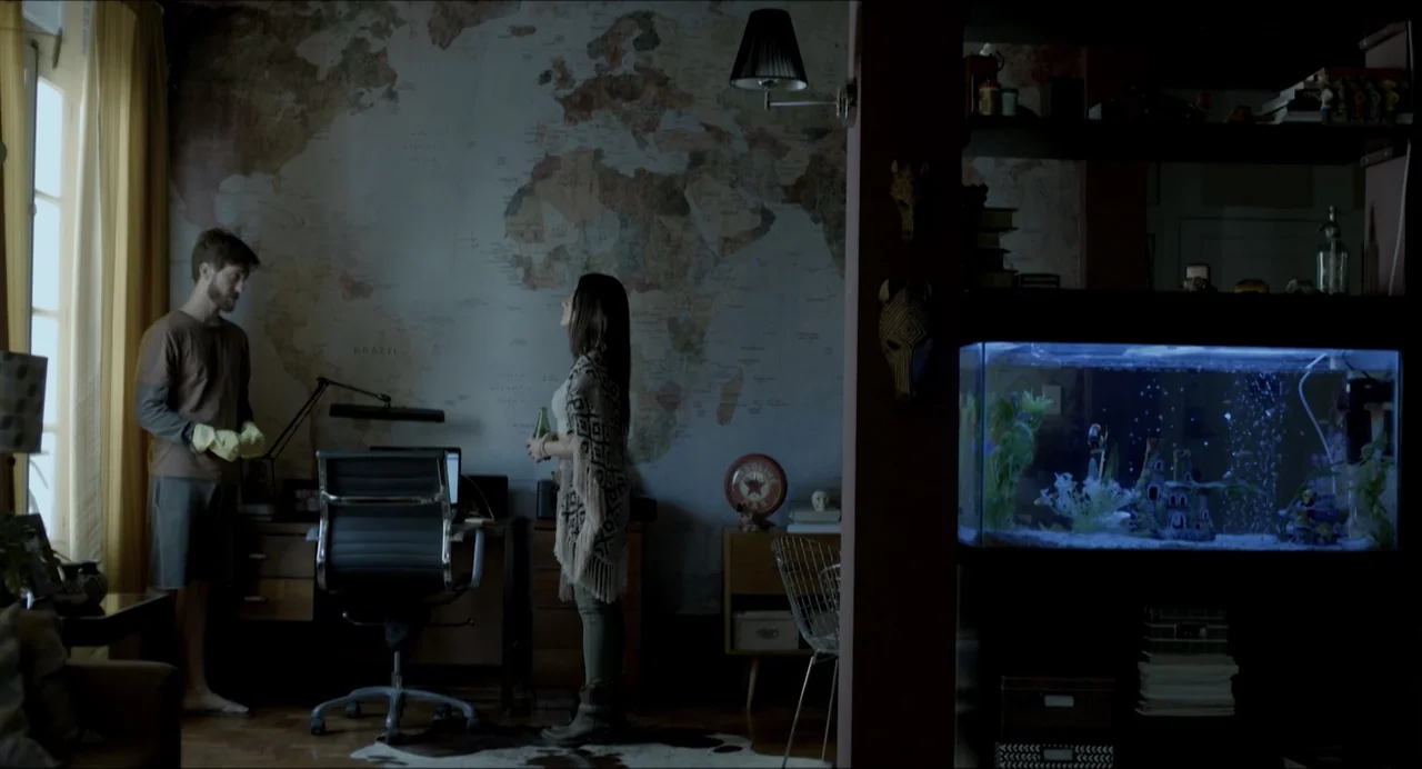 Suspension Points (2022) movie still: fullbody — A furnished residential room features a wall-sized vintage world map and a glass fish tank. A man…