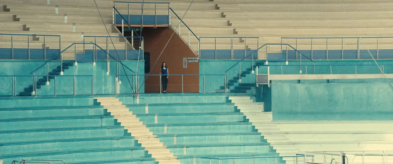 Rust And Bone (2012) movie still: wide — A tiered outdoor stadium features teal and cream-colored seating with metal safety railings. A woman stands…