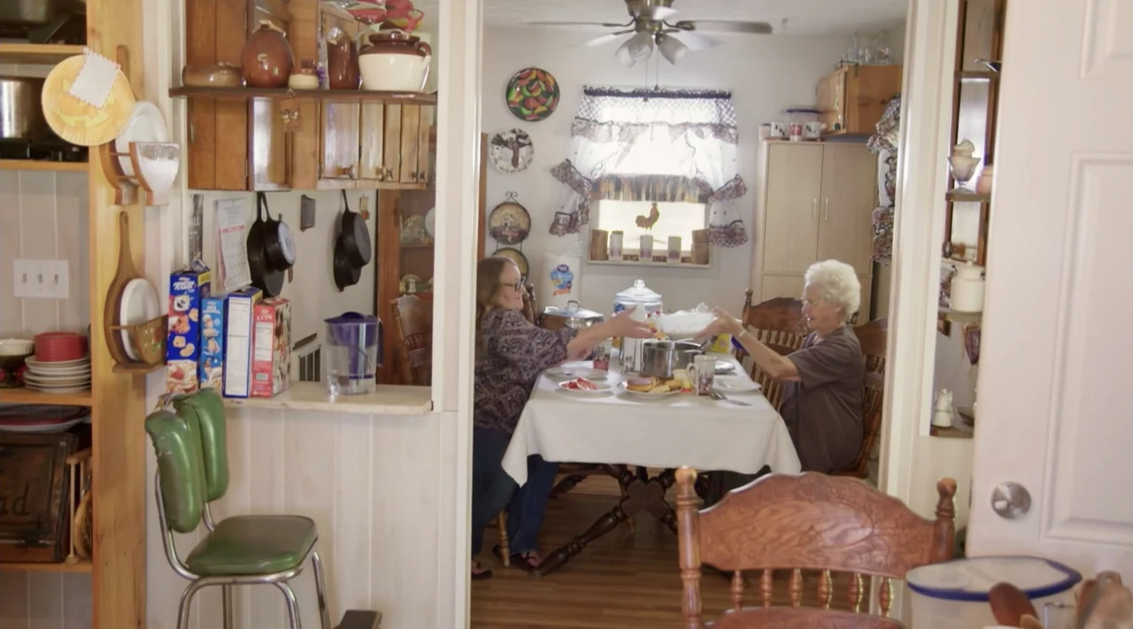The Divided Soul of America (2019) movie still: medium — A cluttered dining area contains a table, chairs, wooden cabinets, and hanging decor. Two…