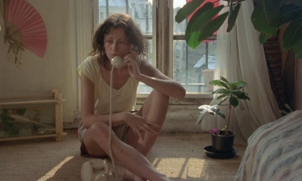 The Green Ray (1986) movie still: medium — A sunlit room with beige carpeting, a potted plant, and a pink wall fan. A woman in a yellow shirt sits…