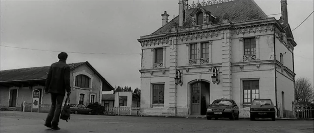 13 Tzameti (2006) movie still: wide — A stone building with ornate architectural detailing sits adjacent to a paved open area. A man walks away from…
