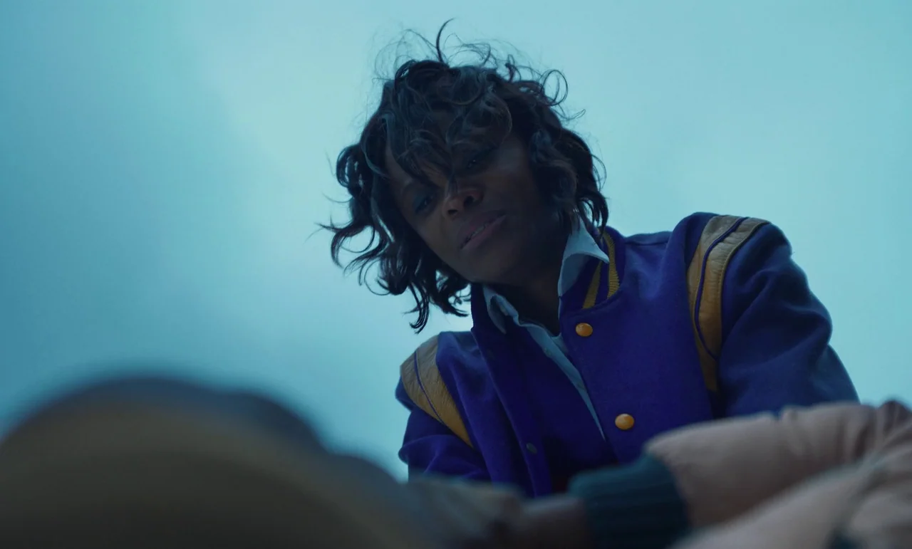 The Silent Twins (2022) movie still: medium — An outdoor pavement area under a cloudy blue-hour sky. A woman wearing a varsity jacket gazes downward…
