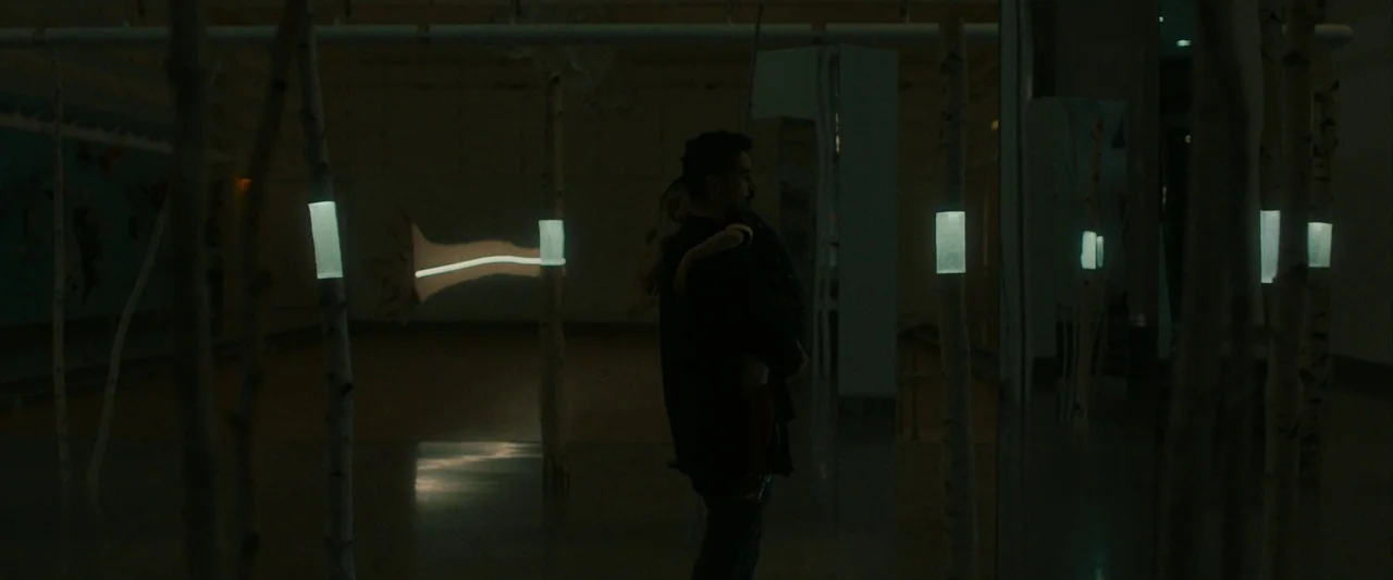 After Yang (2021) movie still: medium — A dark gallery space features several thin birch-like poles equipped with rectangular glowing lights. A man…