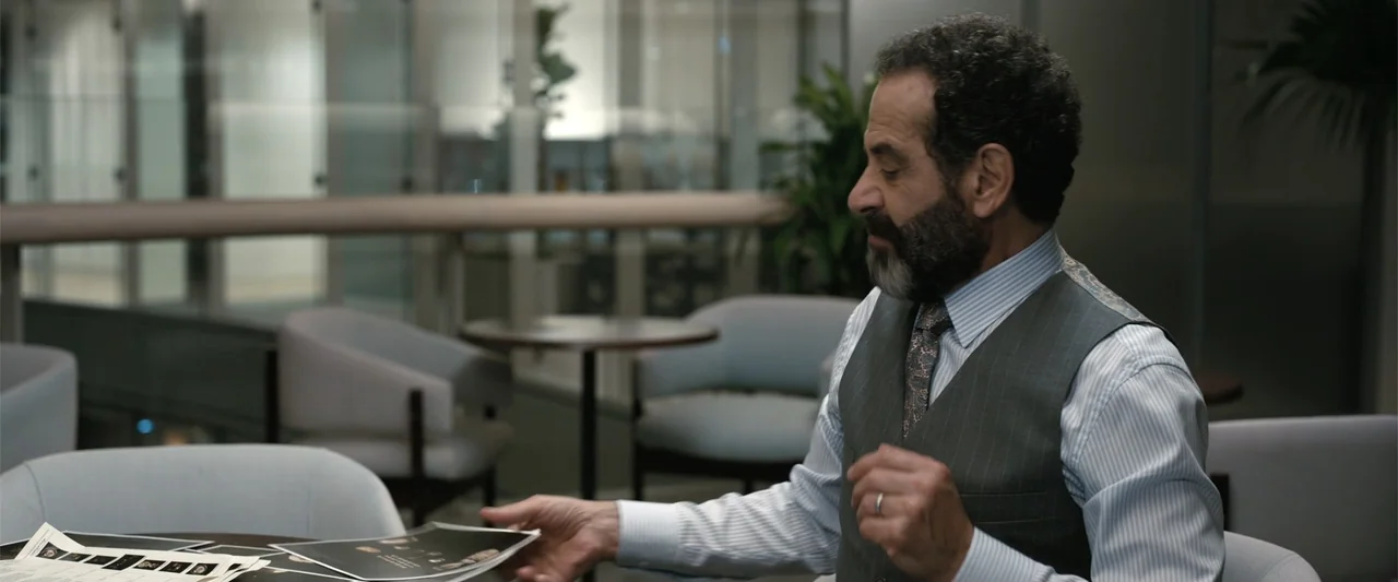 Play Dirty (2025) movie still: medium — A modern lobby with glass partitions and gray armchairs in the background. A middle-aged man in a pinstripe…