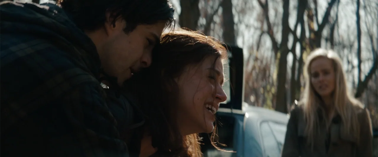 Red Dawn (2012) movie still: medium — A man and woman embrace in a wooded area with bare trees in the background. A third person with blonde hair…